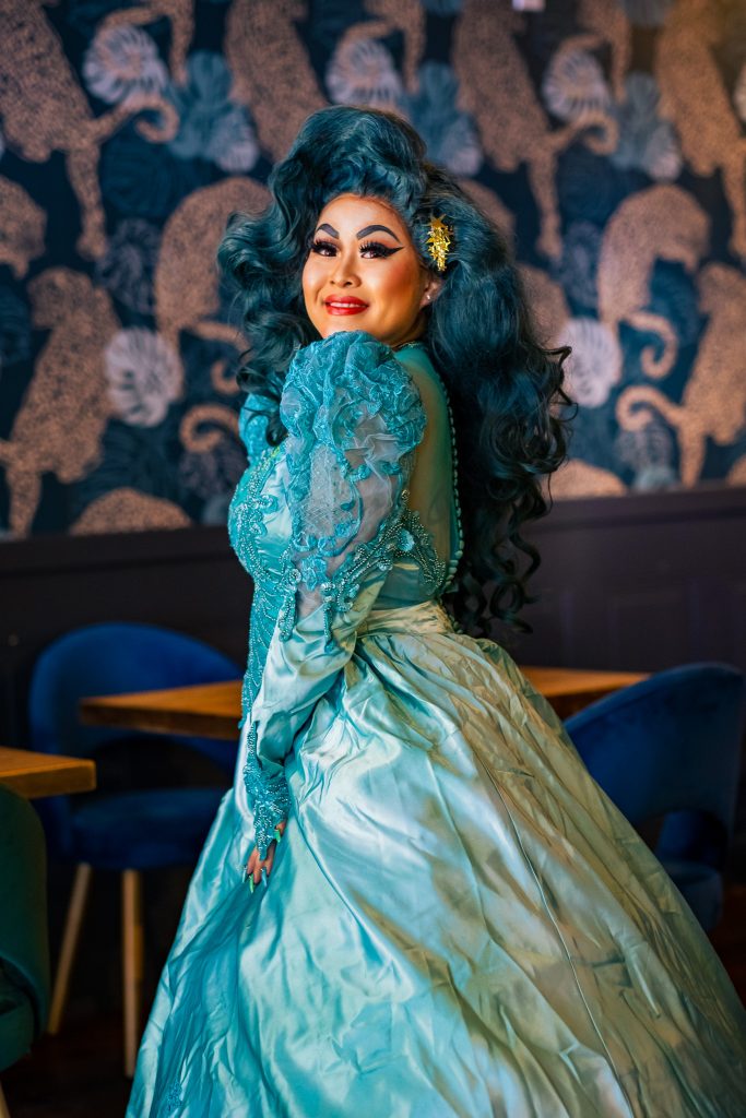 A photo of Hennessy In a teal blue gown with bold makeup and a styled deep teal blue wig.