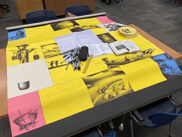 A photo of a large yellow, blue, white, and pink canvas scroll with black line art on it atop a table in the library, along with art materials and books.