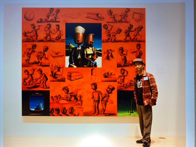 A photo of Evri Kwong standing next to a large work of art hung on a wall.