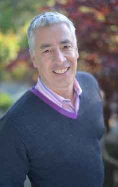 A photo of Michael Genhart smiling at the camera. He has short gray hair and is wearing a blue sweater.